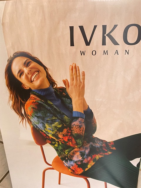 Lachende Frau in bunter Jacke und blauem Pullover, sitzt auf rotem Stuhl vor IVKO WOMAN Plakat.